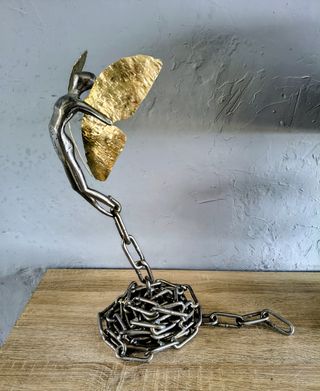 Metal figure reaching towards gold-coloured wing-like forms above a base of tangled steel chains, by Dan'Art.