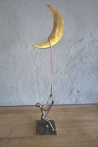 Slender welded metal figure beneath a tall, gold-coloured crescent on a metal stand, by Dan'Art.